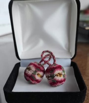 Dos bolitas de lana bordadas con flores en tonos rosa y granate, unidas por un cordón trenzado, dentro de una caja.